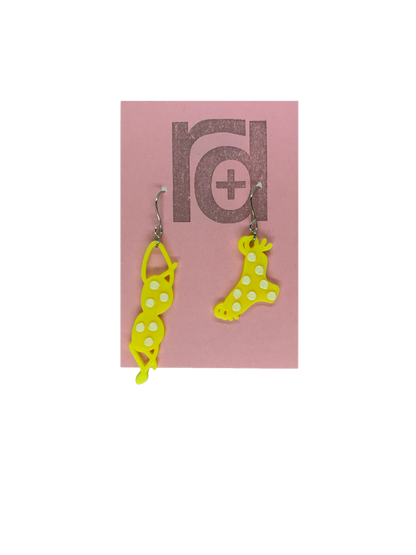 Hanging off of a pink earring card are two R+D earrings. They are asymmetrical  earrings shaped like a classic bikini  that is hung out to dry. The bottoms have ties on the sides. They are yellow with white polka dots to match the classic song, teeny tiny yellow polka dot bikini