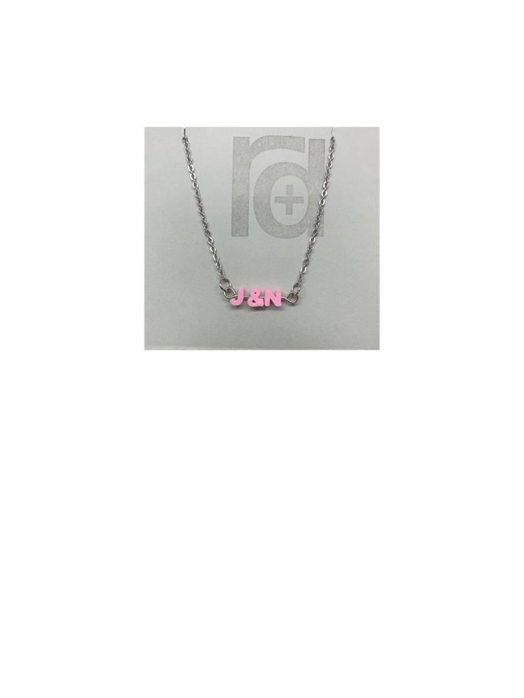 On a grey necklace card is a necklace with small letters on a metal bar. The letters are like beads and will rotate and move around. This necklace has 3 characters (J&N) in a light pink. 