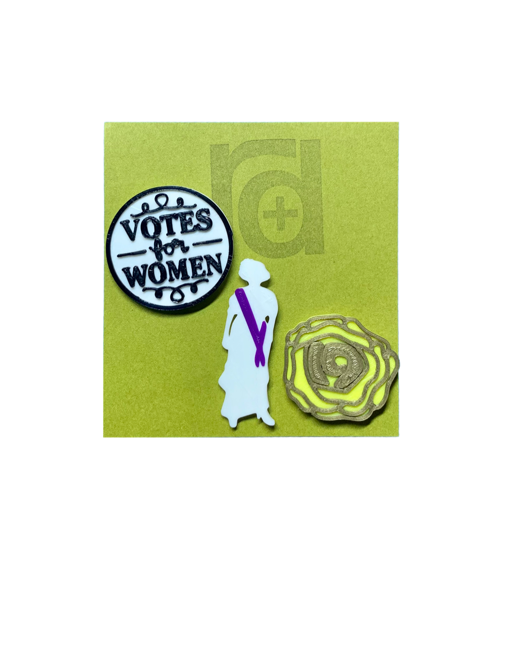 Shown on a green R+D card are a set of three 3D printed pins. The first on the left is a white circle with a black outline and words saying "Votes for Women". The next is a silhouette of a woman from the 1920s. Aside from the silhouette, there is a purple sash. The third pin is a yellow rose, which was worn in support of the movement. The petals are all outlined in gold and in the center is the number 19 for the 19th amendment. 