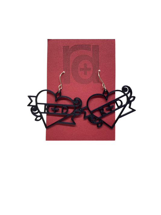 On a dark red earring card are two black R+D earrings. The earrings are shaped as hearts with a banner twisted around it and two small roses. On the banner is reads R+D, which can be personalized to say anything.